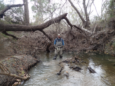 Dr. Christopher Patrick stands in a brown stream in front of several huge downed trees in the mud.