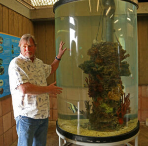 Scott Simon, standing next to a water-filled glass tank with coral