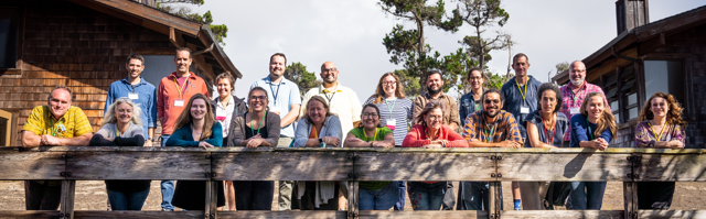 group of about 24 people, leaning over a deck railing in the sunshine